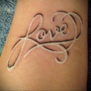 Фото белой татуировки надписью 'Love' в форме сердца на запястье. Минималистичный и изящный рисунок татуировки.