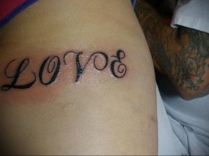 Фотография татуировки надписью 'Love' на боку. Большие буквы, каллиграфический шрифт, женственный дизайн.