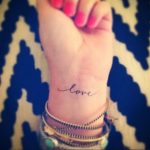 Фотография черной татуировки с надписью 'Love' на запястье. Простой и элегантный шрифт придает татуировке нежный и романтичный вид.