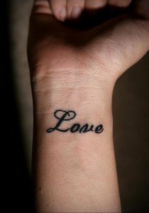 Фотография черной татуировки с надписью 'Love' на запястье. Изящный, рукописный шрифт придает татуировке нежный и романтичный вид.
