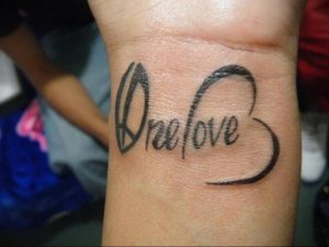 Фото тату на запястье: надпись 'One Love' в каллиграфическом стиле, объединенная с контуром сердца. Простой и элегантный дизайн.