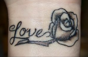 Фото тату на запястье: черная роза с надписью 'love'. Детализированный рисунок, сочетание цветочного мотива и романтической надписи.