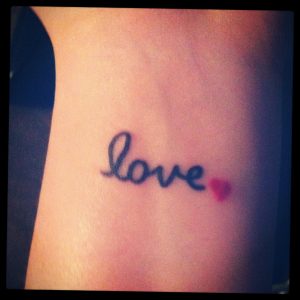 Фотография татуировки на коже с надписью 'Love' и небольшим красным сердцем в конце, выполненной в мягких тонах.