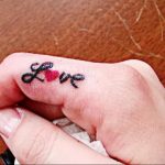 Фотография крошечной татуировки с надписью 'Love' на пальце руки, с красным сердцем вместо буквы 'o'.