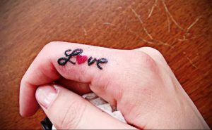 Фотография крошечной татуировки с надписью 'Love' на пальце руки, с красным сердцем вместо буквы 'o'.