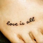 Фото тату на ладони с надписью 'Love is all', выполненной тонким черным шрифтом. Минималистичный дизайн, небольшая татуировка.