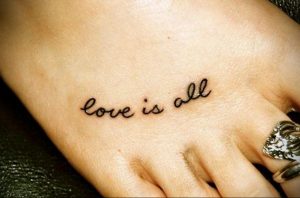 Фото тату на ладони с надписью 'Love is all', выполненной тонким черным шрифтом. Минималистичный дизайн, небольшая татуировка.