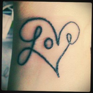 Фото тату на коже с изображением слова 'Love', написанного внутри контура сердца. Черный контур, каллиграфический шрифт.