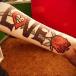 Фото тату на предплечье с надписью 'Love', где буква 'O' заменена на красное сердце, рядом изображена яркая красная роза.