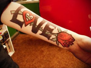 Фото тату на предплечье с надписью 'Love', где буква 'O' заменена на красное сердце, рядом изображена яркая красная роза.