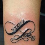 Фото тату бесконечность с римскими цифрами 'V.XXIV.X' и надписью 'love' на предплечье. Черно-белый рисунок татуировки в минималистичном стиле.