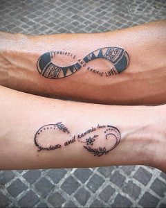 Фото парных татуировок бесконечность на предплечьях с надписью 'symbiotic and karmic love' и цветами, выполненные в черно-серых тонах.