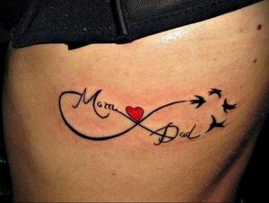 Фото тату бесконечность на бедрах с надписями 'Mom' и 'Dad' и тремя летящими птицами, выполненное в черных тонах с красным сердцем.