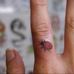 Фото тату маленькой красной божьей коровки на пальце. Простой рисунок татуировки, минималистичный дизайн, небольшой размер.