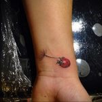 Фото милой татуировки божьей коровки, сидящей на одуванчике, на запястье. Изображение выполнено в ярких, реалистичных цветах.