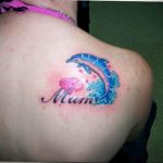 Фото тату дельфина с надписью 'Mum' и изображением сердца. Татуировка выполнена в ярких цветах и расположена на плече. Трогательный рисунок татуировки.