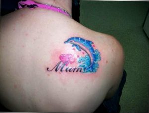 Фото тату дельфина с надписью 'Mum' и изображением сердца. Татуировка выполнена в ярких цветах и расположена на плече. Трогательный рисунок татуировки.