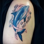 Красивое фото тату дельфина, выпрыгивающего из воды, с надписью "Your battle is our battle" и розовой лентой. Татуировка выполнена в ярких цветах.