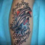 Красочное фото тату дельфина, выпрыгивающего из волн, с надписью "In Loving Memory Mom". Татуировка выполнена в реалистичном стиле с использованием ярких цветов.