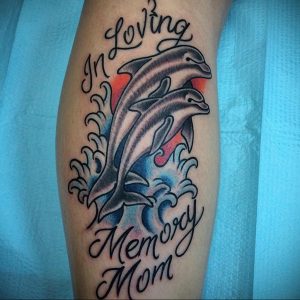 Красочное фото тату дельфина, выпрыгивающего из волн, с надписью "In Loving Memory Mom". Татуировка выполнена в реалистичном стиле с использованием ярких цветов.