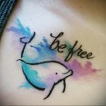 Фото тату дельфина в акварельном стиле с надписью 'Be Free'. Татуировка выполнена в пастельных тонах с использованием голубого, розового и фиолетового.