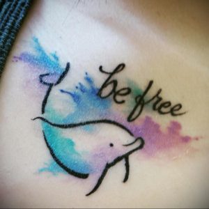 Фото тату дельфина в акварельном стиле с надписью 'Be Free'. Татуировка выполнена в пастельных тонах с использованием голубого, розового и фиолетового.
