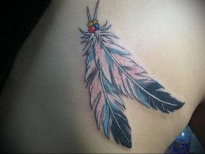 Фото тату: красочное перо в стиле Native American, украшенное бусинами, расположено на боку. Яркие цвета и этнические мотивы.