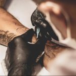 Фото процесса нанесения татуировки: мастер в черных перчатках работает машинкой над татуировкой на руке клиента. Видна часть уже выполненной работы.