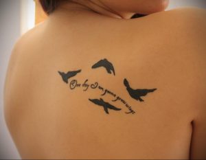 Фото тату: Небольшие черные силуэты летящих птиц с надписью "One day I am gonna grow wings", расположенные на спине.