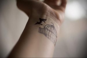 Фото тату: небольшая черно-белая татуировка птицы, вылетающей из клетки, на женском запястье. Простой и элегантный рисунок татуировки.
