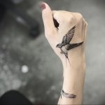 Фото тату: реалистичный рисунок летящей птицы на руке, выполненный в черно-серых тонах. Детализированная татуировка с проработкой перьев.