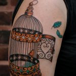 Фото тату с изображением открытой клетки и вылетающих птиц на плече. Детализированный рисунок в стиле олд скул с использованием ярких цветов.