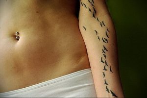 Фото тату: стая птиц, летящих вдоль руки. Минималистичный и элегантный рисунок татуировки, выполненный в черном цвете.