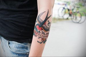 Фото тату ласточки в стиле олд скул на предплечье. Яркие цвета, четкие контуры, надпись 'FREEBIRD'. Идеи для тату, эскизы татуировок.