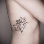 Фото тату колибри на боку, выполненное в черно-белых тонах. Изящный рисунок птицы среди нежных цветов.