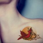 Фото тату красного кардинала на ветке с желтым ореолом на ключице девушки. Красивый рисунок татуировки в ярких цветах.
