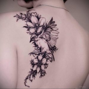 Фото тату ласточки, окруженной цветами, на спине, выполненной в технике реалистичной графики с использованием черного и серого цветов.