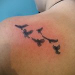 Несколько маленьких черных татуировок птиц, летящих в одну сторону на спине девушки. Минималистичный и элегантный рисунок татуировки.