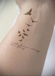 Фото тату: стая птиц, взлетающих вверх по запястью, с надписью 'With brave wings she flies'. Элегантный и мотивирующий рисунок.