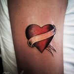 Фото тату: классическая татуировка сердца с лентой, обвивающей его, и стрелой, пронзающей сердце. Расположена на плече.