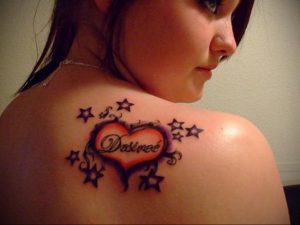 Фото тату: небольшое сердце с именем 'Desiree' внутри, окруженное звездами. Татуировка расположена на спине девушки.