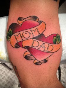 Фото тату: классическая татуировка сердца с надписью 'Mom Dad' на ленте, выполненная в традиционном стиле. Яркий и памятный рисунок татуировки.