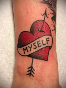 Фото тату красного сердца, пронзенного стрелой, с надписью 'MYSELF' на ленте. Олдскульный стиль татуировки на руке.