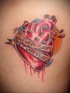 Фото тату: Красное сердце с надписью "Never Say Forever" на ленте, пронзенное стрелами и с капающей кровью. Драматичный и эмоциональный рисунок татуировки на боку.