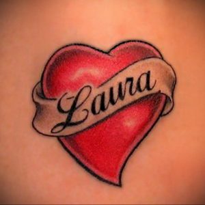 Фото тату: Красное сердце с именем "Laura" на белой ленте. Классический и романтичный рисунок татуировки на коже.
