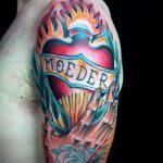 Фото татуировки на плече: сердце в огне, с надписью "Moeder" на ленте. Яркий и драматичный рисунок в стиле олд скул.