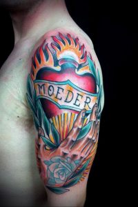 Фото татуировки на плече: сердце в огне, с надписью "Moeder" на ленте. Яркий и драматичный рисунок в стиле олд скул.