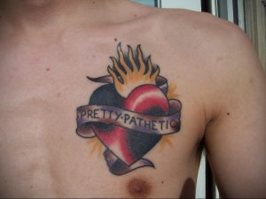 Фото тату горящего сердца с надписью 'Pretty Pathetic' на груди. Яркий и драматичный рисунок татуировки, символизирующий боль, страсть и разочарование.