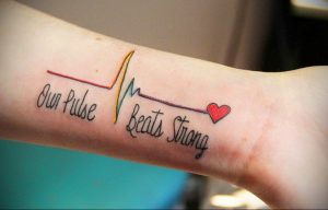 Фото тату с изображением пульса, надписью 'Our Pulse Beats Strong' и маленьким красным сердцем на руке. Символичный рисунок татуировки, посвященный любви и жизни.