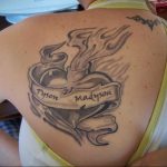 Фото тату: черно-серое тату горящего сердца с именами 'Tyson' и 'Madyson' на ленте. Татуировка на спине.